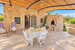Un espace de restauration en plein air avec un mur en pierre, une table en bois, des chaises blanches et un toit de chaume.