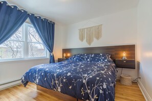 Ein Schlafzimmer mit einem Bett, einem Fenster mit Vorhängen, einem Nachttisch mit Lampe und einem Wandbehang aus Makramee.