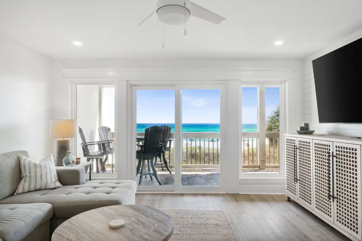 A living room with a sofa, a coffee table, and a TV on a cabinet. There is a balcony with a view of the ocean.