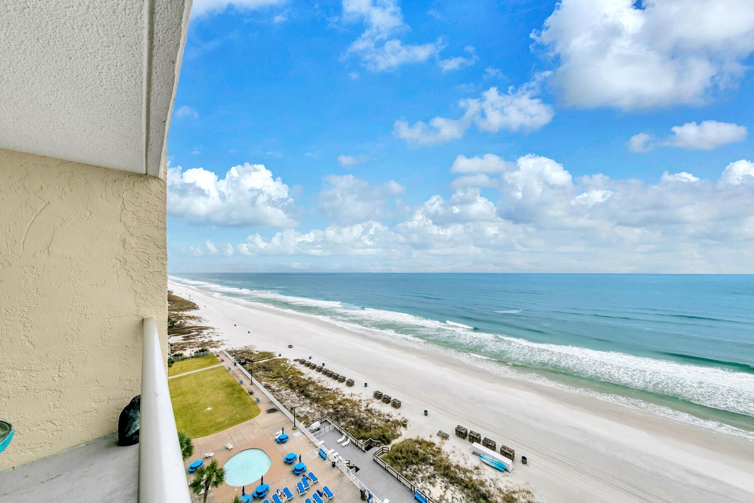 A balcony with a view of a beach, a pool area with lounge chairs, and a clear sky.