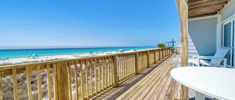 A wooden deck with a view of the beach, a white table, and a blue umbrella.