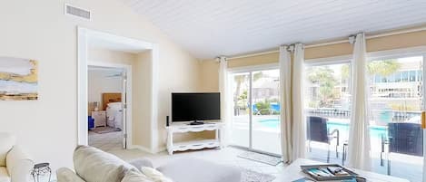 A living room with a sofa, a coffee table, and a TV. There is a door leading to a balcony with a pool view.