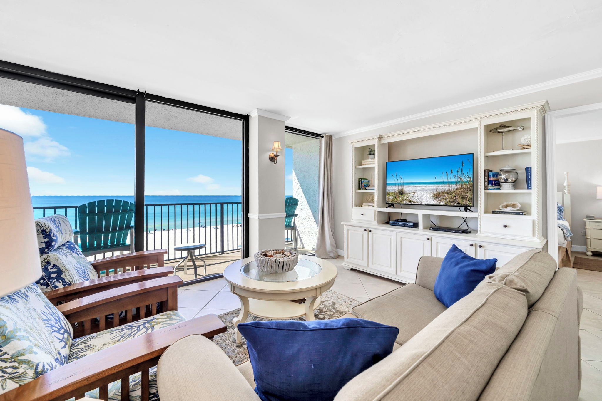 A living room with a large window overlooking the beach, a sofa, a coffee table, and a TV on a white cabinet.