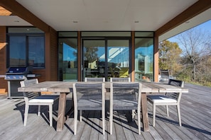 Een terras buiten met een houten dek, een barbecue en een tafel met stoelen.