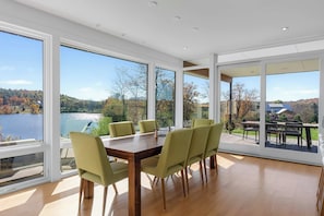 Een eetgedeelte met een tafel en stoelen, grote ramen met uitzicht op een meer en bomen, en een terras met buitenmeubilair.