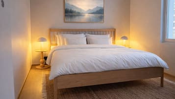 Un dormitorio con una cama de madera, ropa de cama blanca, dos lámparas de noche y un cuadro con un paisaje en la pared.