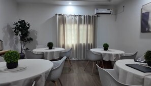 Ein Hotelzimmer mit vier runden Tischen, jeder mit einer weißen Tischdecke und einer Topfpflanze. Es gibt ein Fenster mit Vorhängen, eine Klimaanlage an der Wand und ein gerahmtes Bild.