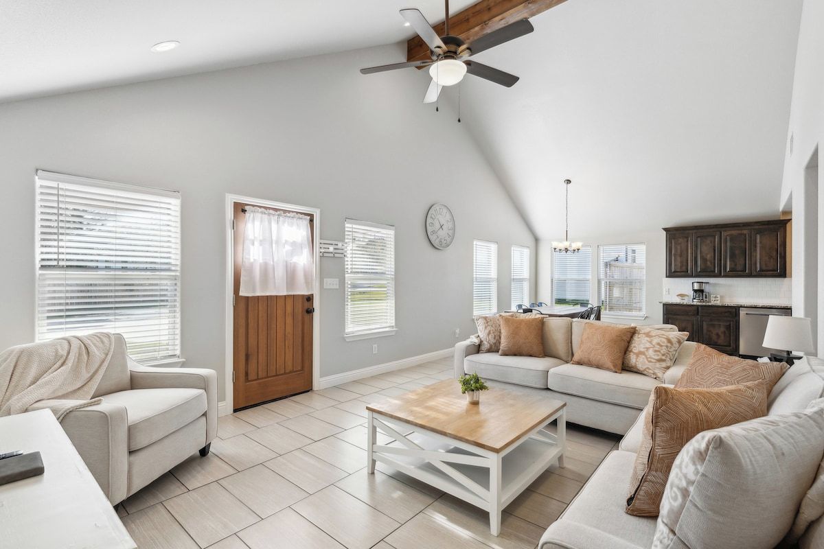 A spacious living area with a ceiling fan, a sofa set, a coffee table, and a kitchen area in the background.