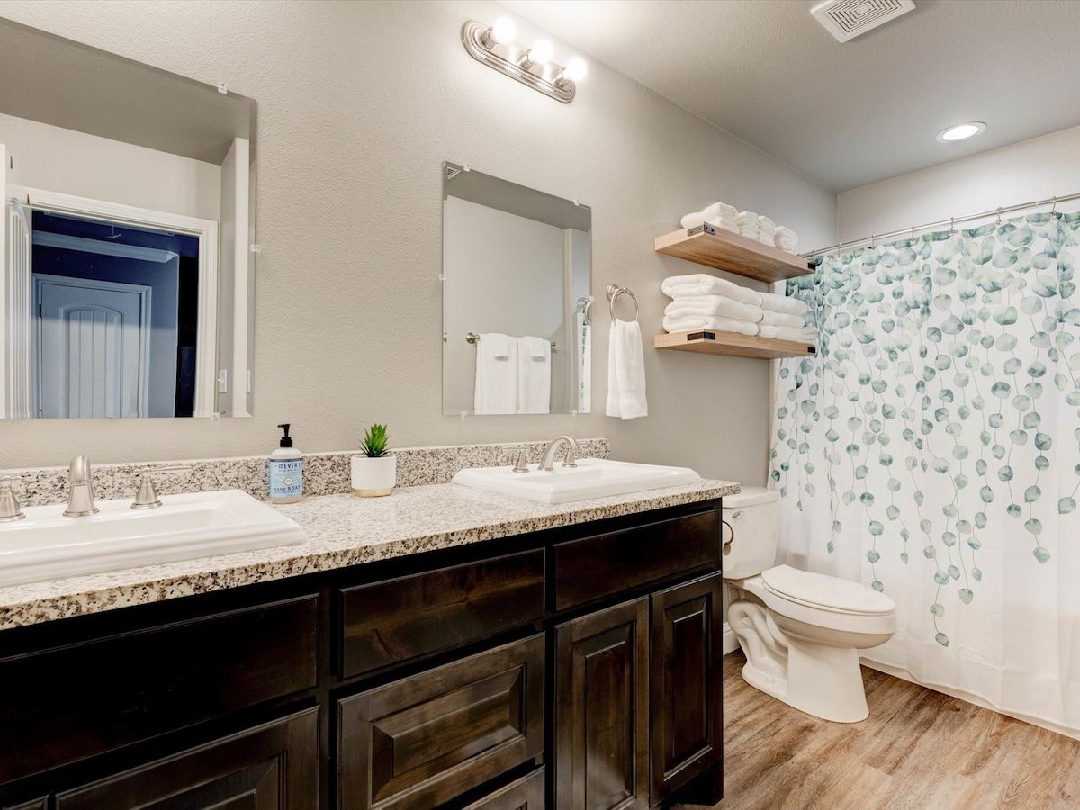 A bathroom with a double sink, a toilet, a shower curtain with a leaf pattern, and a mirror with lights above.
