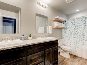 A bathroom with a double sink, a toilet, a shower curtain with a leaf pattern, and a mirror with lights above. - Cozy 2 Bedroom Apartment in Historic Granbury (Granbury)