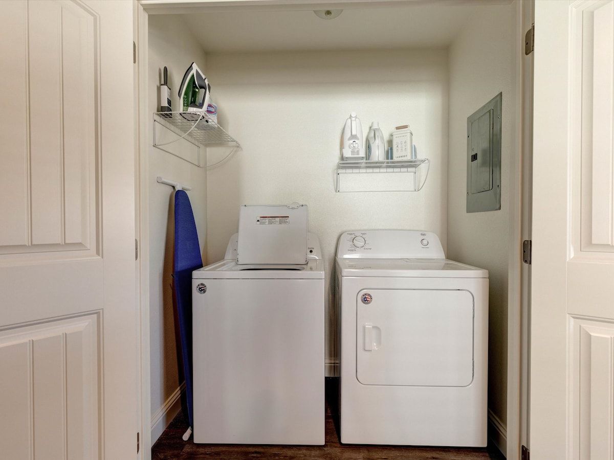 A laundry room with two white washing machines and a dryer, a shelf with cleaning supplies, and a blue towel hanging on the wall.