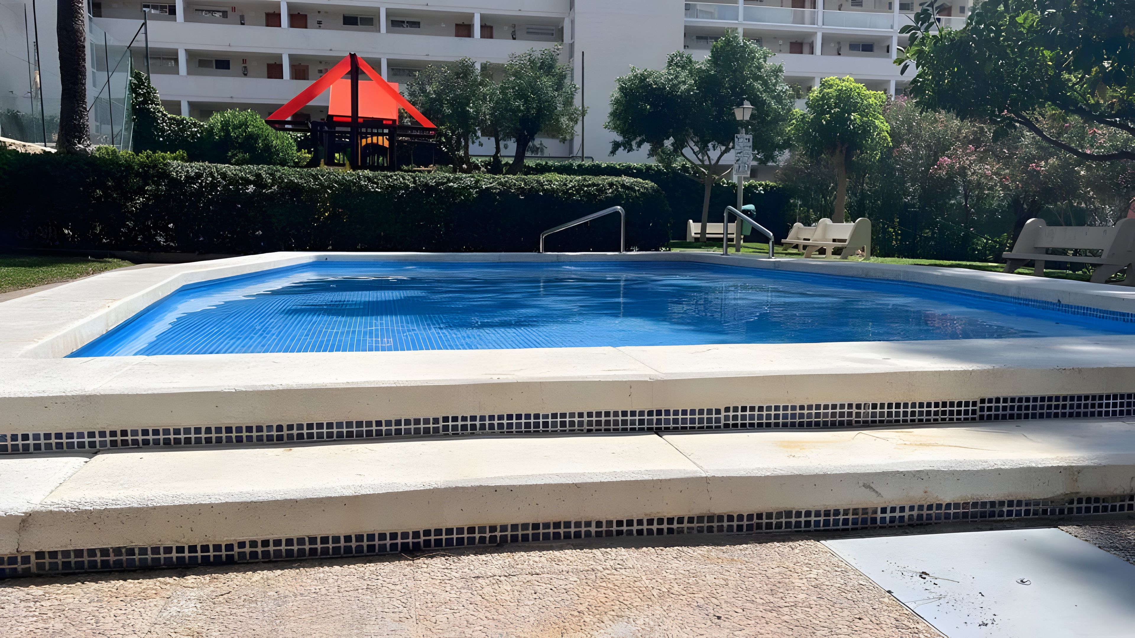 Una piscina amb vora de rajola, envoltada per una zona pavimentada, i un edifici amb balcons al fons.