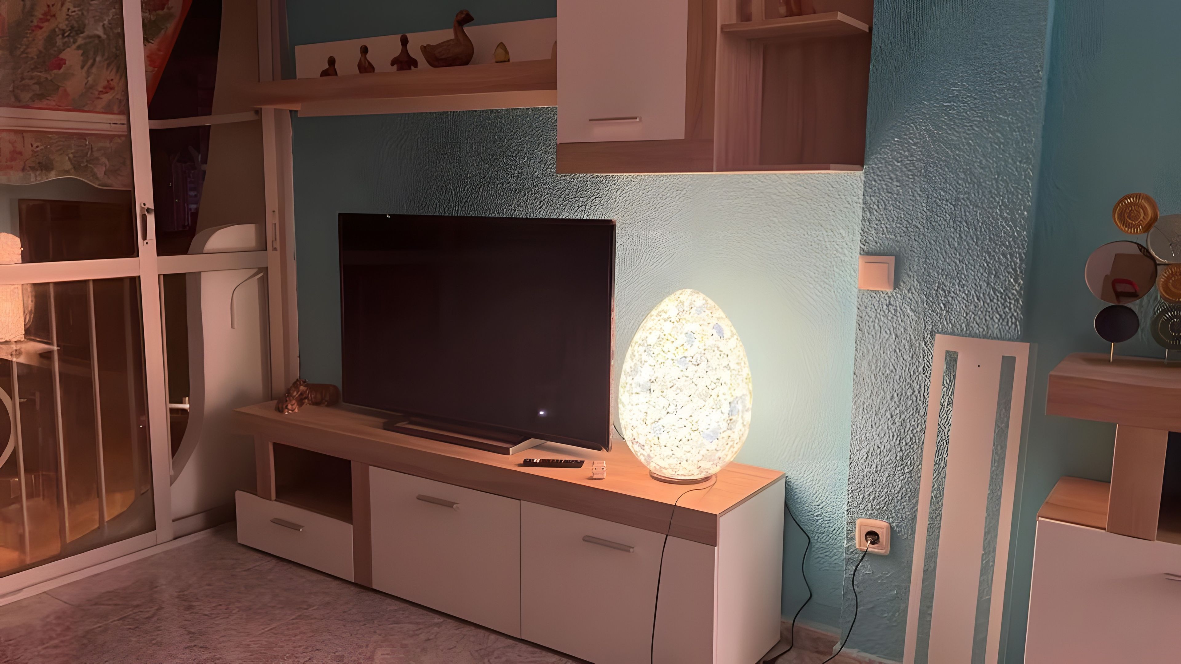 Quarto com televisão sobre suporte de madeira, luminária decorativa e parede com texturas.