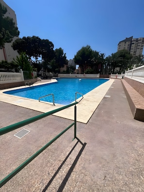 Una piscina con superficie de agua azul cristalina, rodeada por una valla blanca baja, y contigua a un edificio de techo plano.