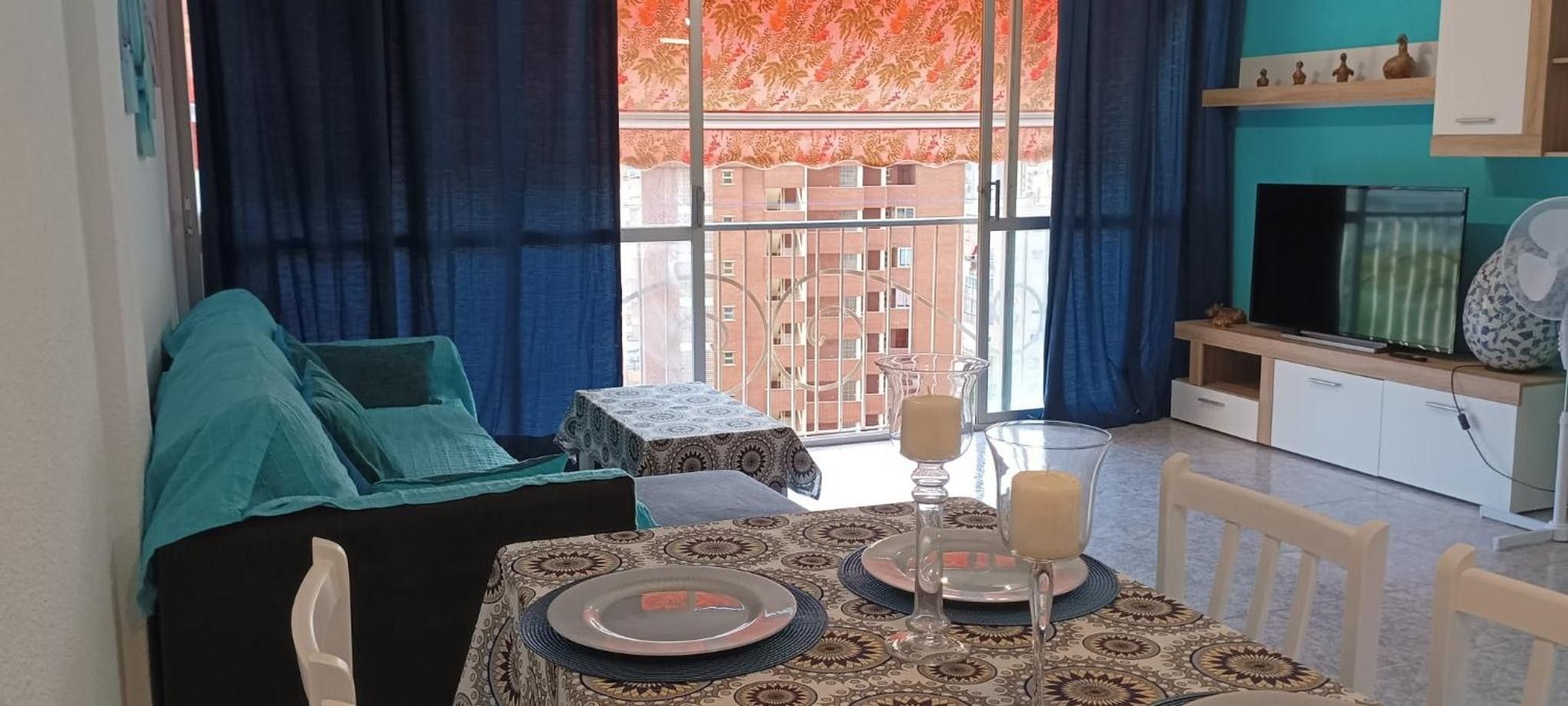 Uma zona de refeições com uma mesa posta para dois, um sofá e vista para um edifício através de uma janela.