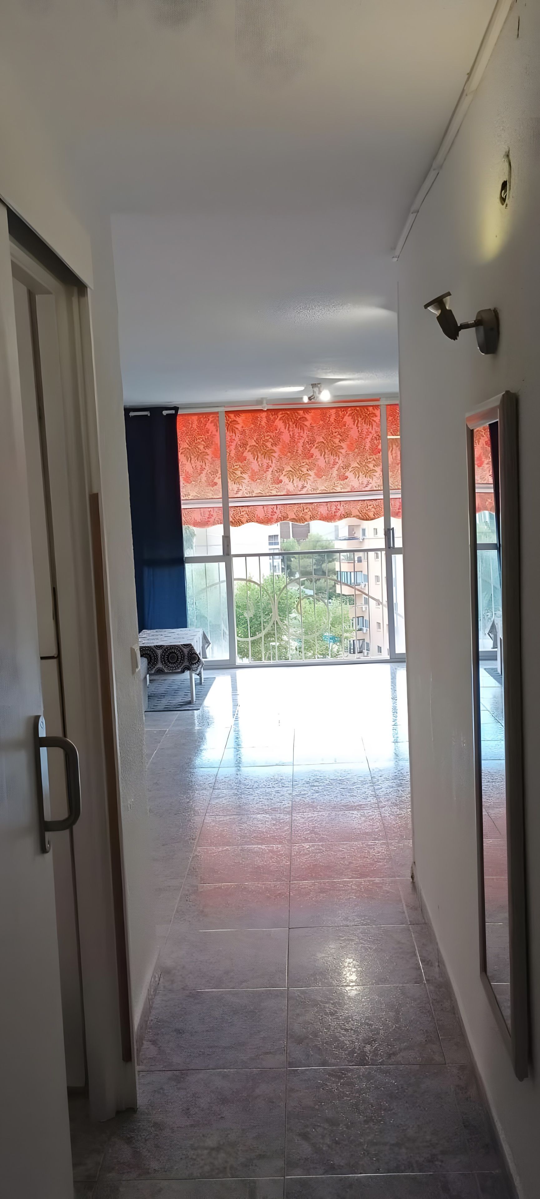 Um corredor com piso ladrilhado que leva a uma varanda com vista para edifícios e árvores.