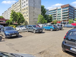 Parkkipaikka, jossa on useita eri automerkkejä, korkea rakennus ja monikerroksinen rakennus, jossa on suuret ikkunat.