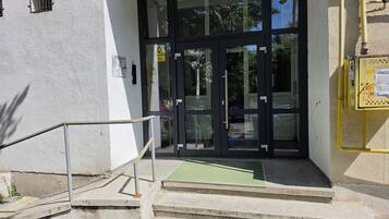 Entrada de un edificio con puerta de vidrio, barandilla metálica y un cuadro eléctrico amarillo.