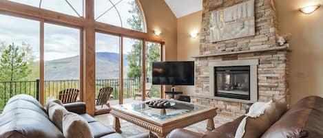 A spacious living area with a stone fireplace, leather sofas, a flat-screen TV, and a large window offering a mountain view.