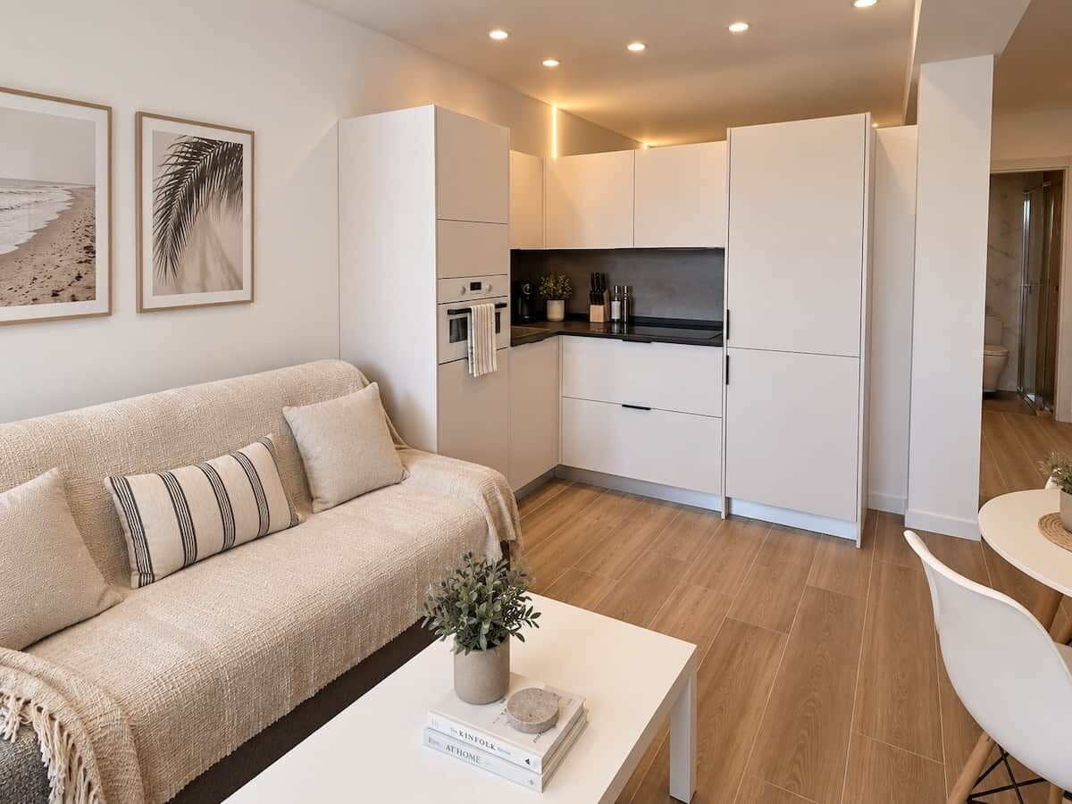Een moderne woonkamer met een bank, een salontafel met een boek en een plant, en een keuken met witte kasten en een aanrecht.