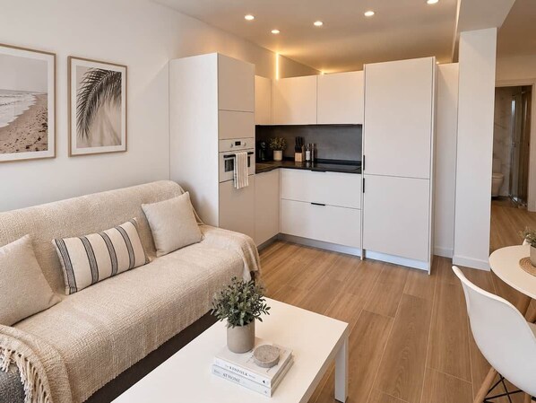Een moderne woonkamer met een bank, een salontafel met een boek en een plant, en een keuken met witte kasten en een aanrecht.