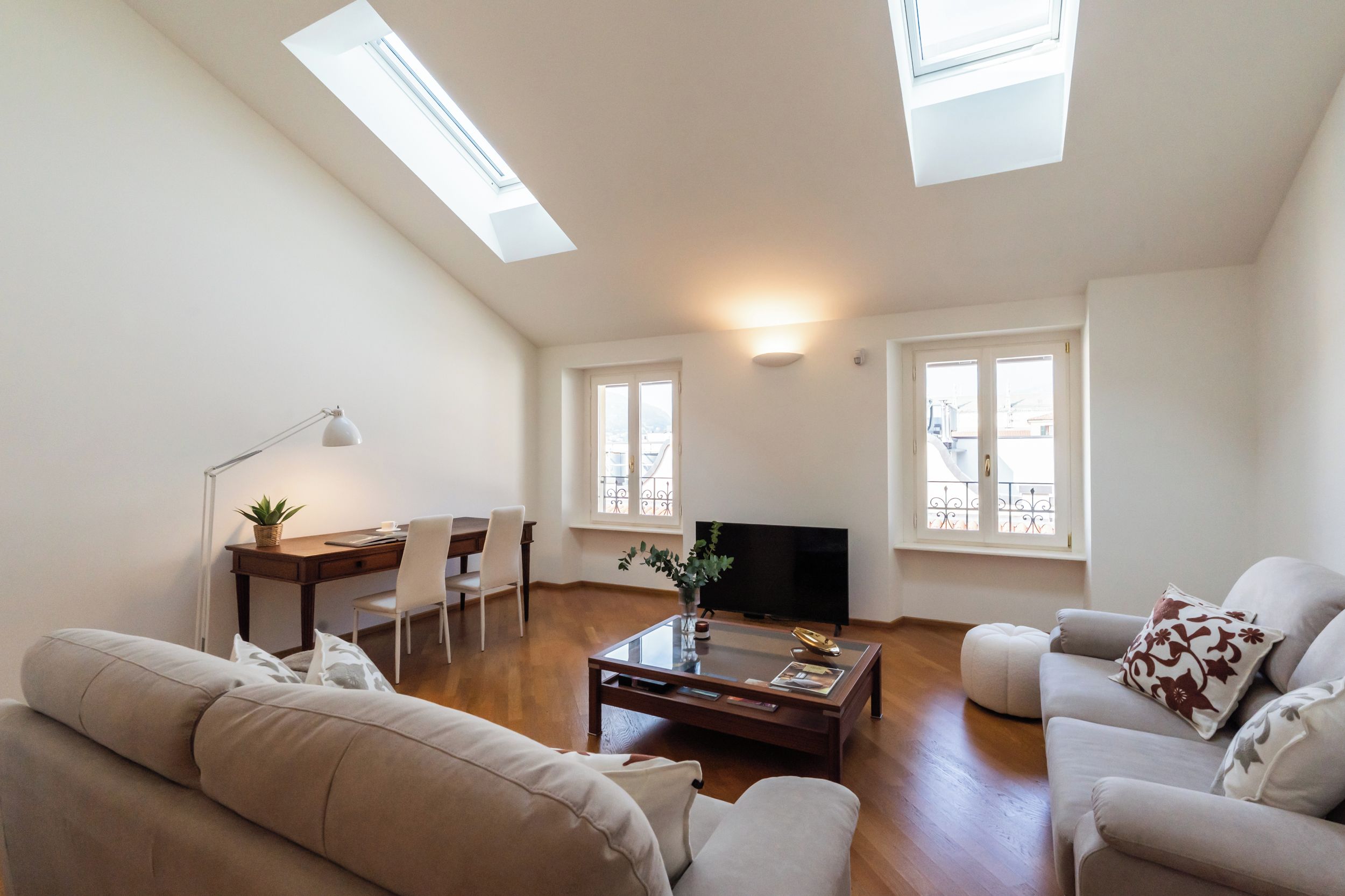 Un salon spacieux comprenant un canapé, une table basse, un téléviseur et un bureau avec des chaises. Des lucarnes sont aménagées au plafond.
