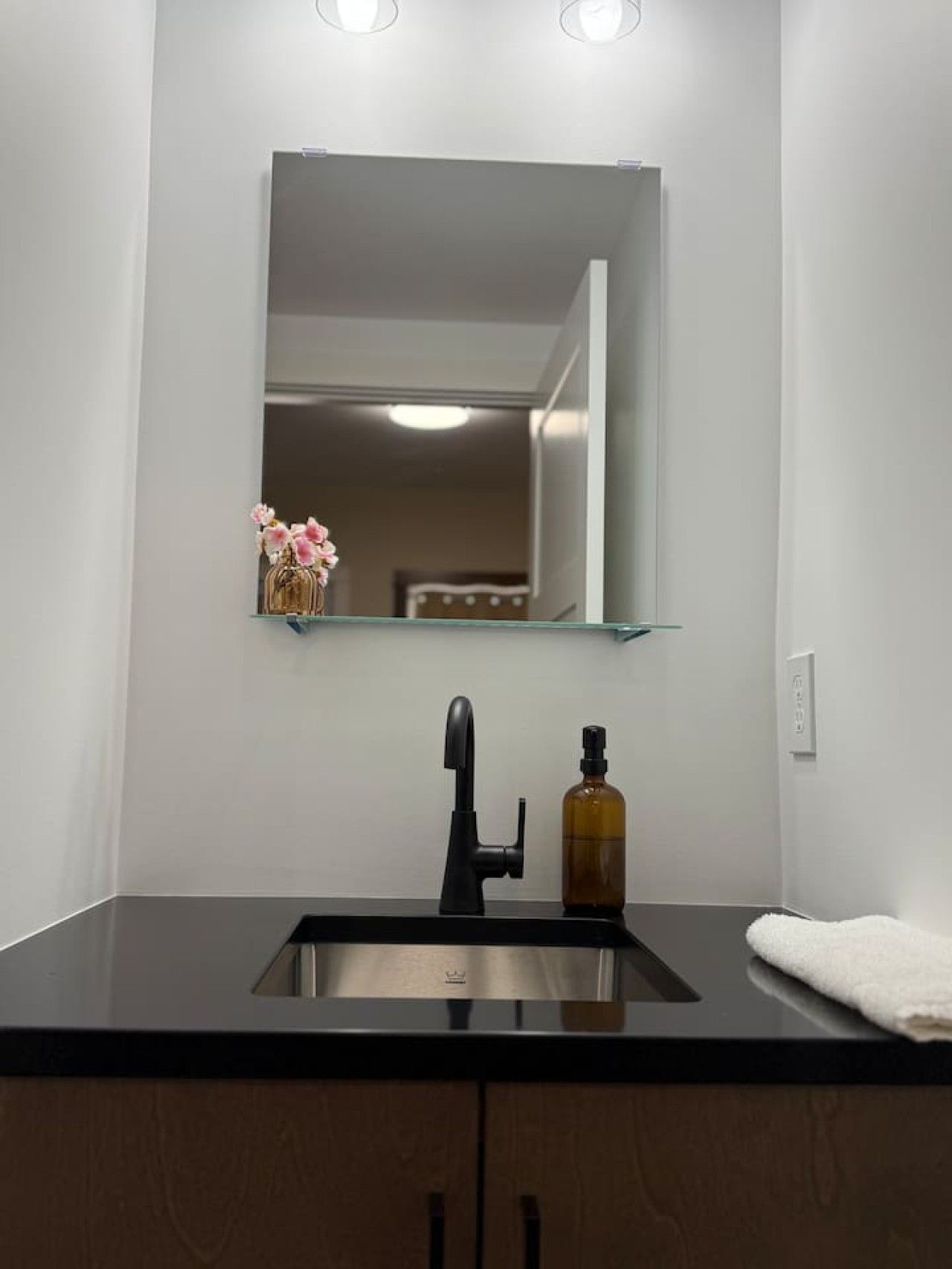 A bathroom with a black countertop, a stainless steel sink, a mirror, and a bottle of hand soap.