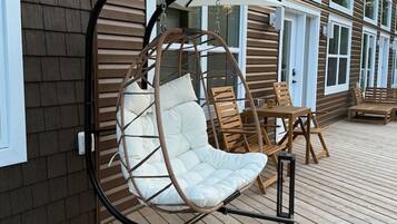Une terrasse en bois avec un fauteuil suspendu et une table, un bâtiment doté de grandes fenêtres et une porte.