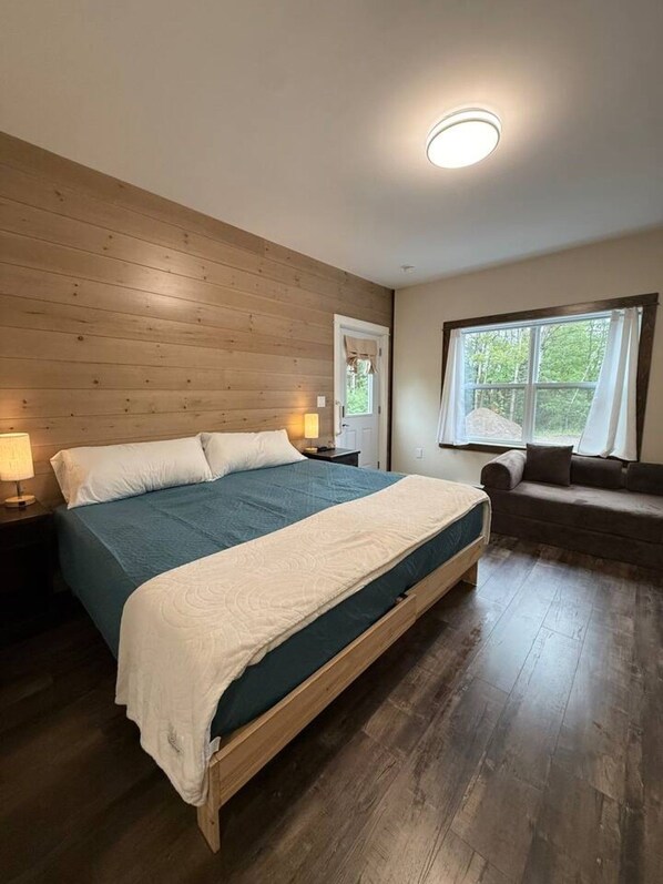 A bedroom with a wooden bed, a nightstand, a window with curtains, and a view of greenery outside. - Sakura Valley Studio Suite 10 | Private Entrance (North Alton)