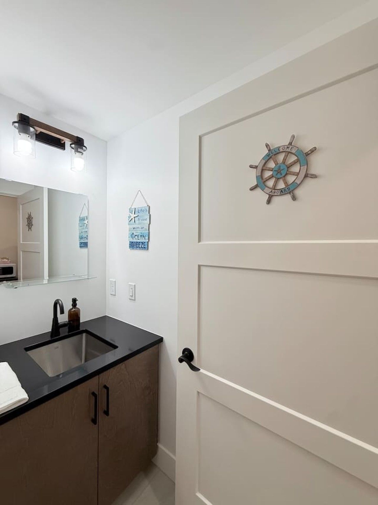 Une salle de bain avec un lavabo noir, un miroir et une porte ornée d’un motif représentant un gouvernail de navire.