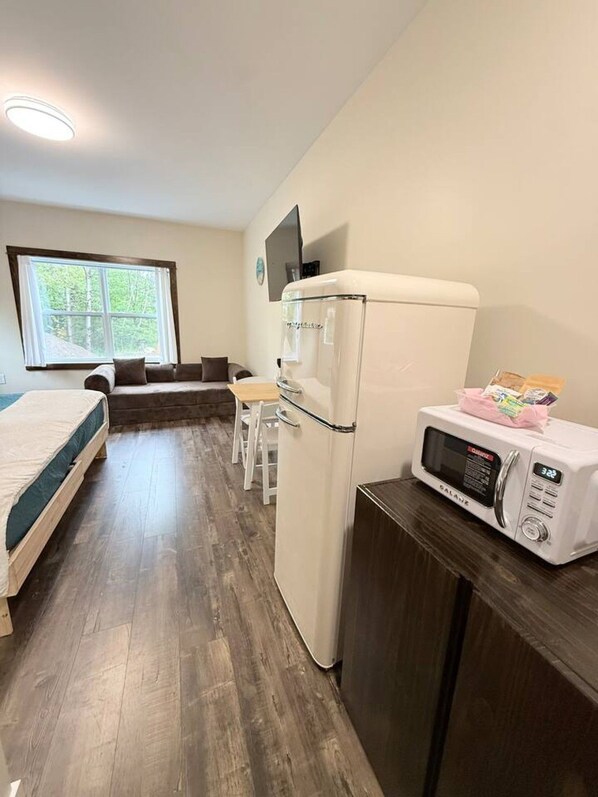 Ett kompakt hotellrum med en säng, ett litet pentry, en liten matplats och ett fönster med utsikt över träd.