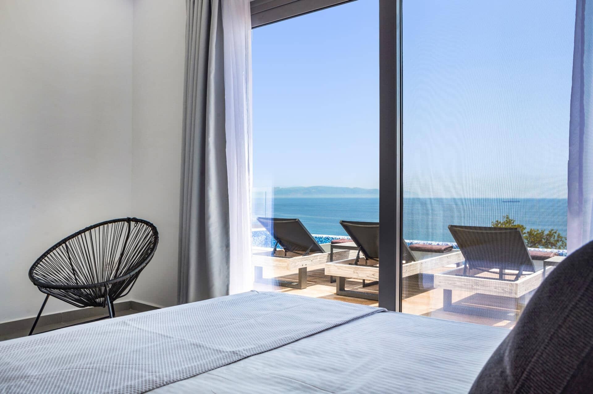 Un dormitorio con un ventanal grande con vista a la playa. Hay una cama con edredón blanco, una silla negra y vista al mar.