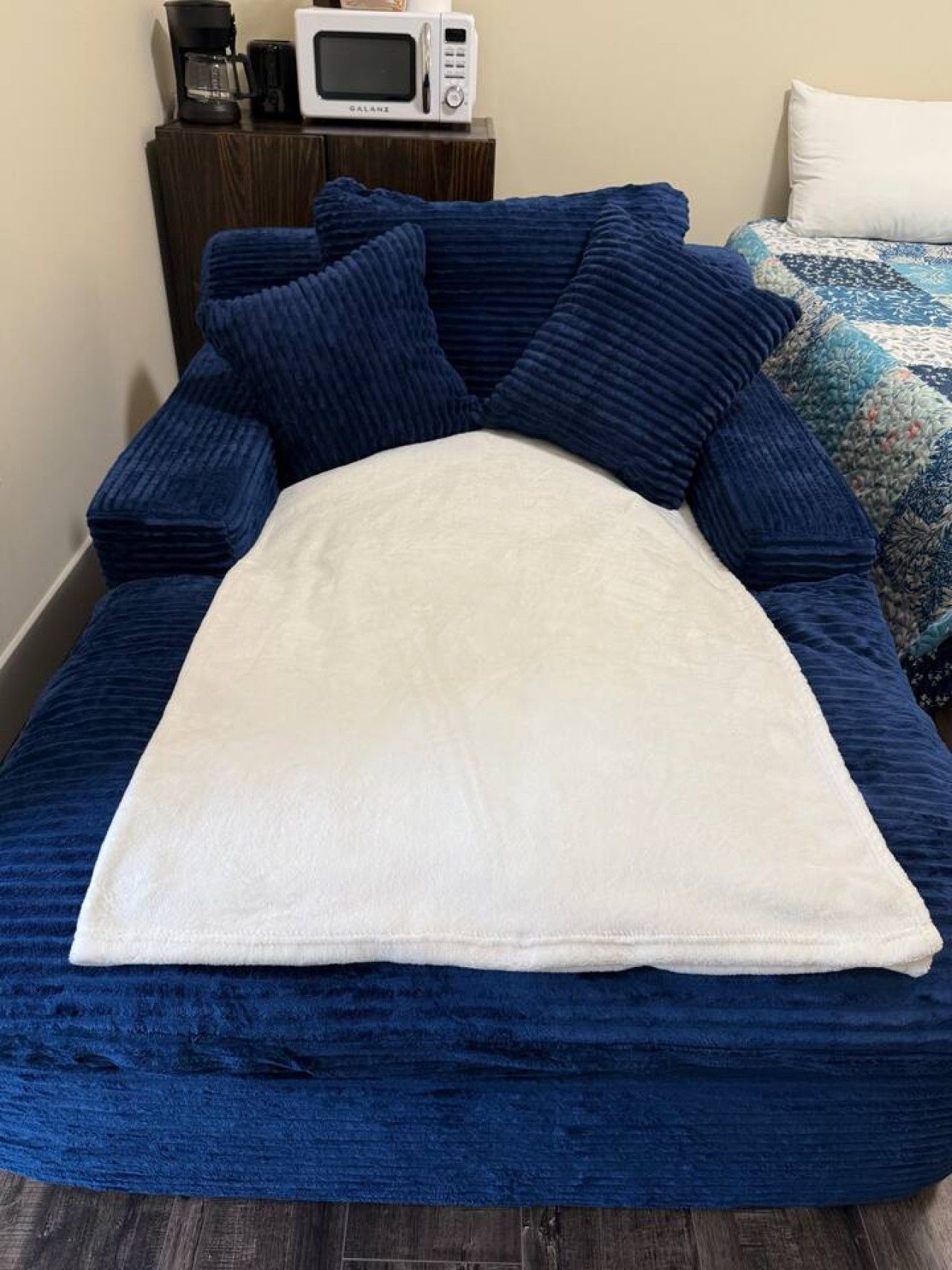 Una cama con una manta blanca, almohadas azules y un colcha. Un microondas y una cafetera están sobre una mesita de noche junto a la cama.