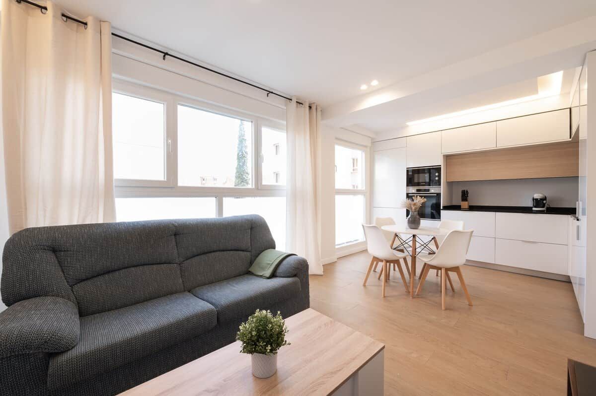 Un salon moderne avec un canapé gris, une petite table ornée d’une plante et un coin repas avec des chaises et une table blanches.