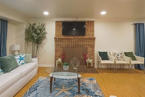A living room with a brick fireplace, a flat-screen TV mounted above it, a glass coffee table, a white sofa with patterned cushions, and a blue floral rug. - Spacious 5-bedroom house in delightful Fountain Valley with AC (Fountain Valley)