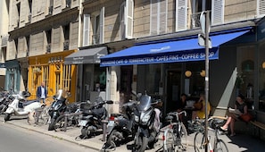 Une rue bordée de motos et de vélos garés, un café avec un auvent bleu et un immeuble dont la devanture est jaune.