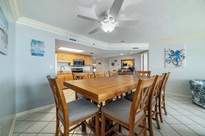 A dining area with a wooden table and chairs, a ceiling fan, and a kitchen in the background. - Phoenix VI #913 - New! Beachfront 3BR, Gulf Views, Indoor/Outdoor Pools, Hot Tub (Orange Beach)