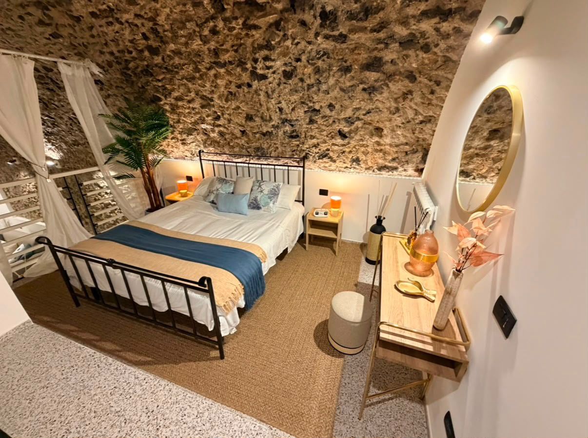 A bedroom with a stone wall, a bed with a blue bedspread, a bedside table with a lamp, and a small stool.