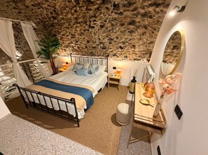 Une chambre avec un mur en pierre, un lit recouvert d’une couverture bleue, une table de chevet avec une lampe et un petit tabouret.
