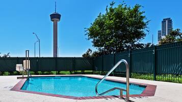 Ein Schwimmbecken mit einer Metallleiter, umgeben von einem grünen Zaun, mit einem hohen Turm und Gebäuden im Hintergrund.