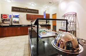 Un espace buffet pour le petit-déjeuner comprenant un distributeur de Coca-Cola, un panneau indiquant que le petit-déjeuner est gratuit et servi quotidiennement, ainsi qu’une variété d’options alimentaires.