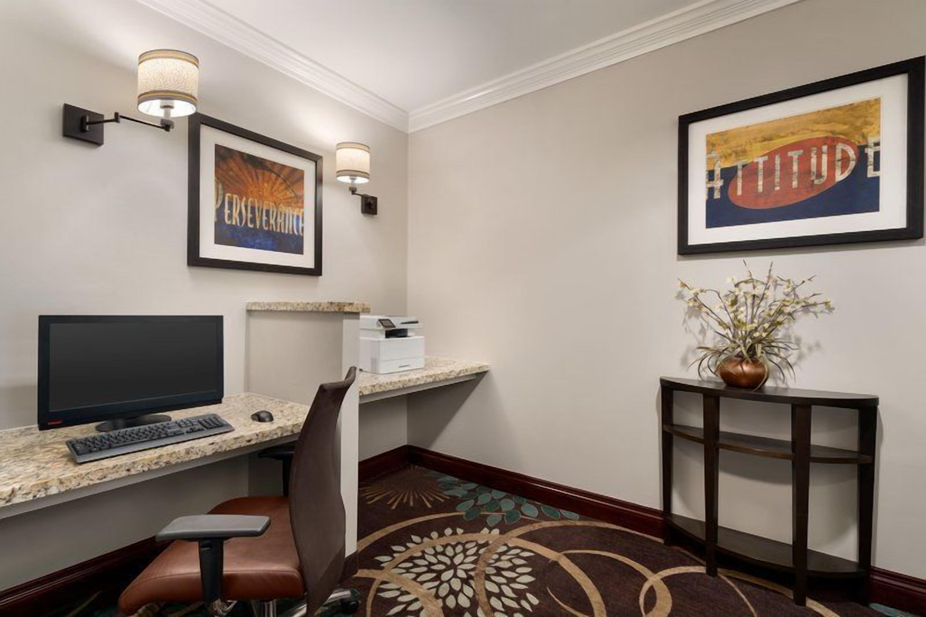 A hotel room with a desk, computer, chair, framed artwork, and a decorative plant.