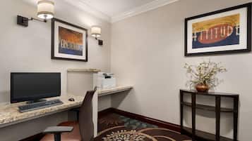 A hotel room with a desk, computer, chair, framed artwork, and a decorative plant.