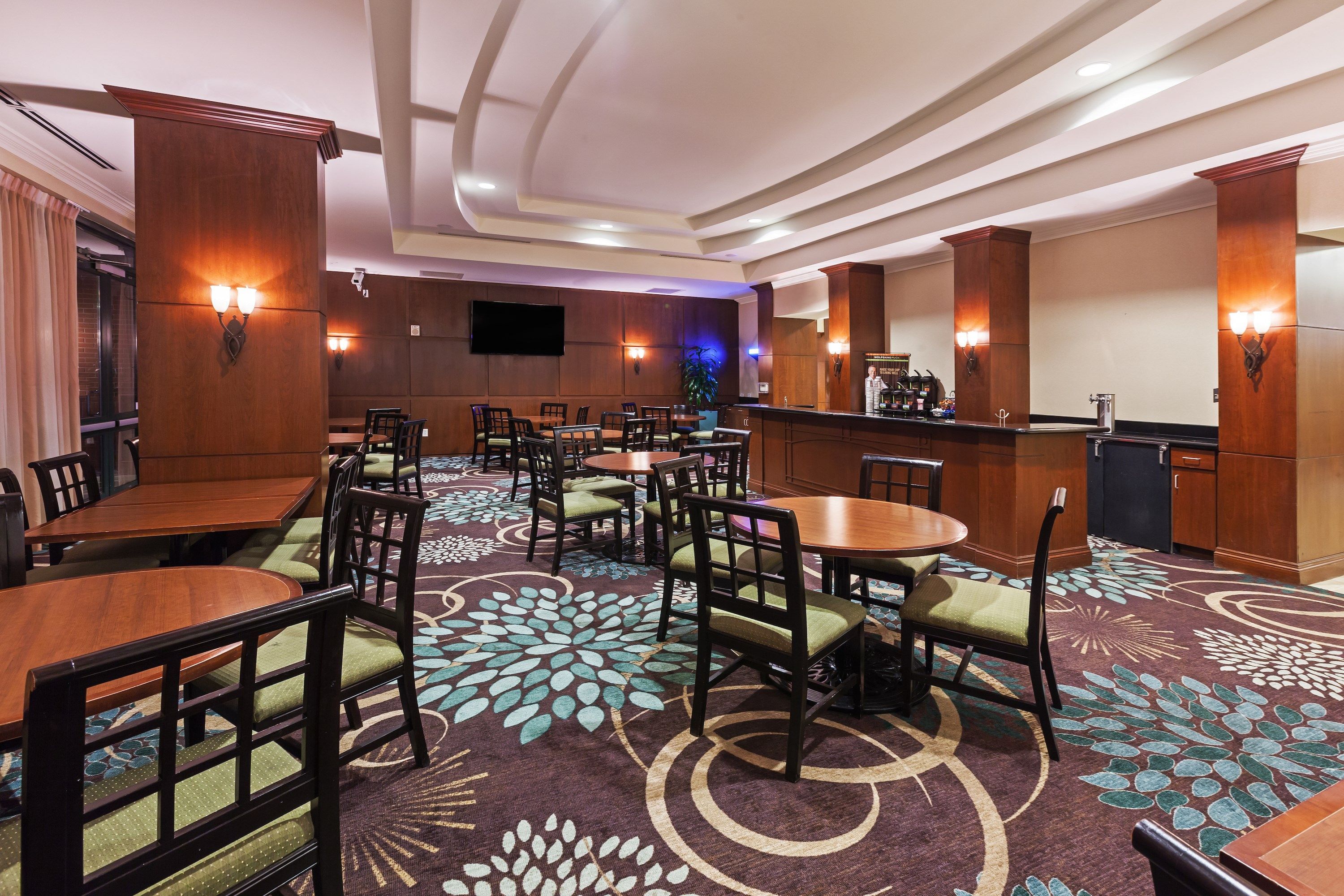 Un vestíbulo de hotel con varias mesas y sillas de comedor, una zona de bar y una alfombra estampada.