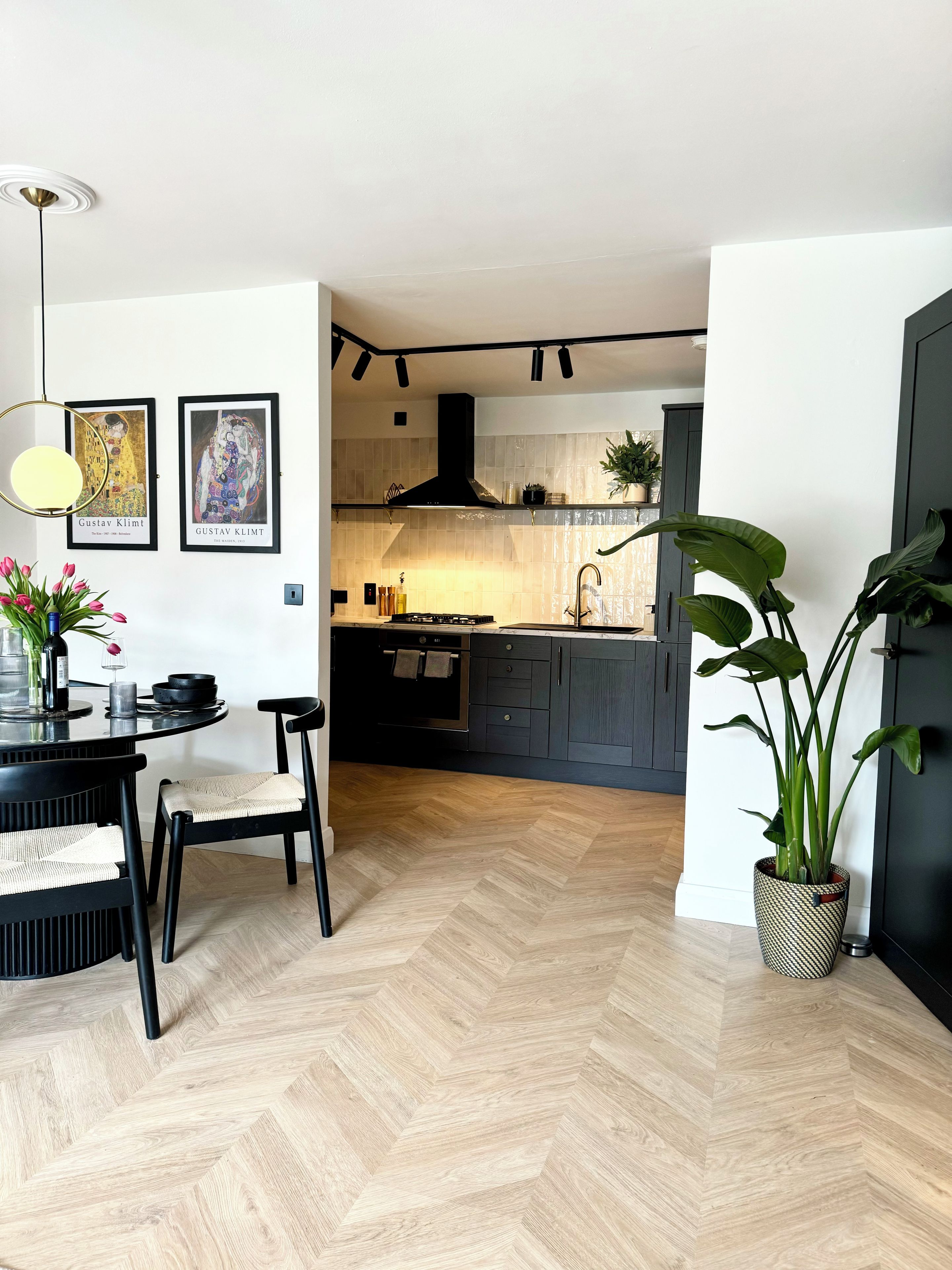 A modern kitchen with a herringbone floor, a dining area with a round table and chairs, and two framed artworks on the wall.