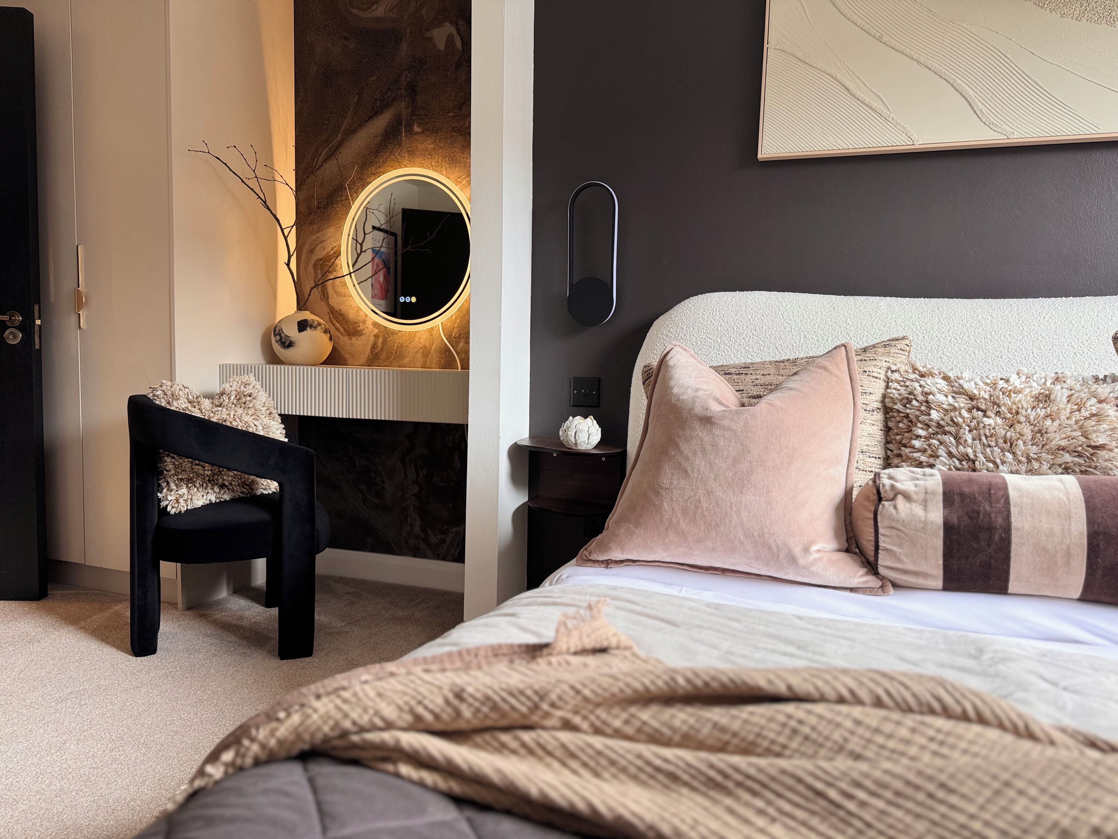 A bedroom with a bed, a chair, a vanity with a mirror, and a wall-mounted light.