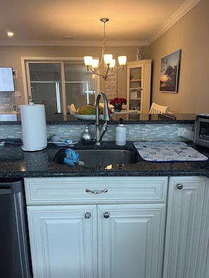 A kitchen with a stainless steel sink, a chandelier, and a view of the living area. - ENJOY THE COMFORTS OF HOME IN THIS MODERN 6 ROOM CONDO. 1/4 BLOCK TO BEACH! (Seaside Park)