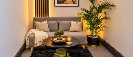 A cozy living room with a sofa, a coffee table, a bonsai tree, a potted plant, and a framed picture on the wall.