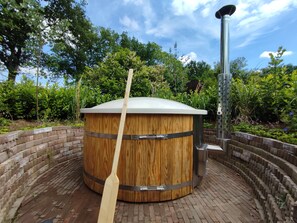 Ein hölzernes Whirlpool mit einem Metallbrenner, umgeben von einer Ziegelwand und viel Grün.