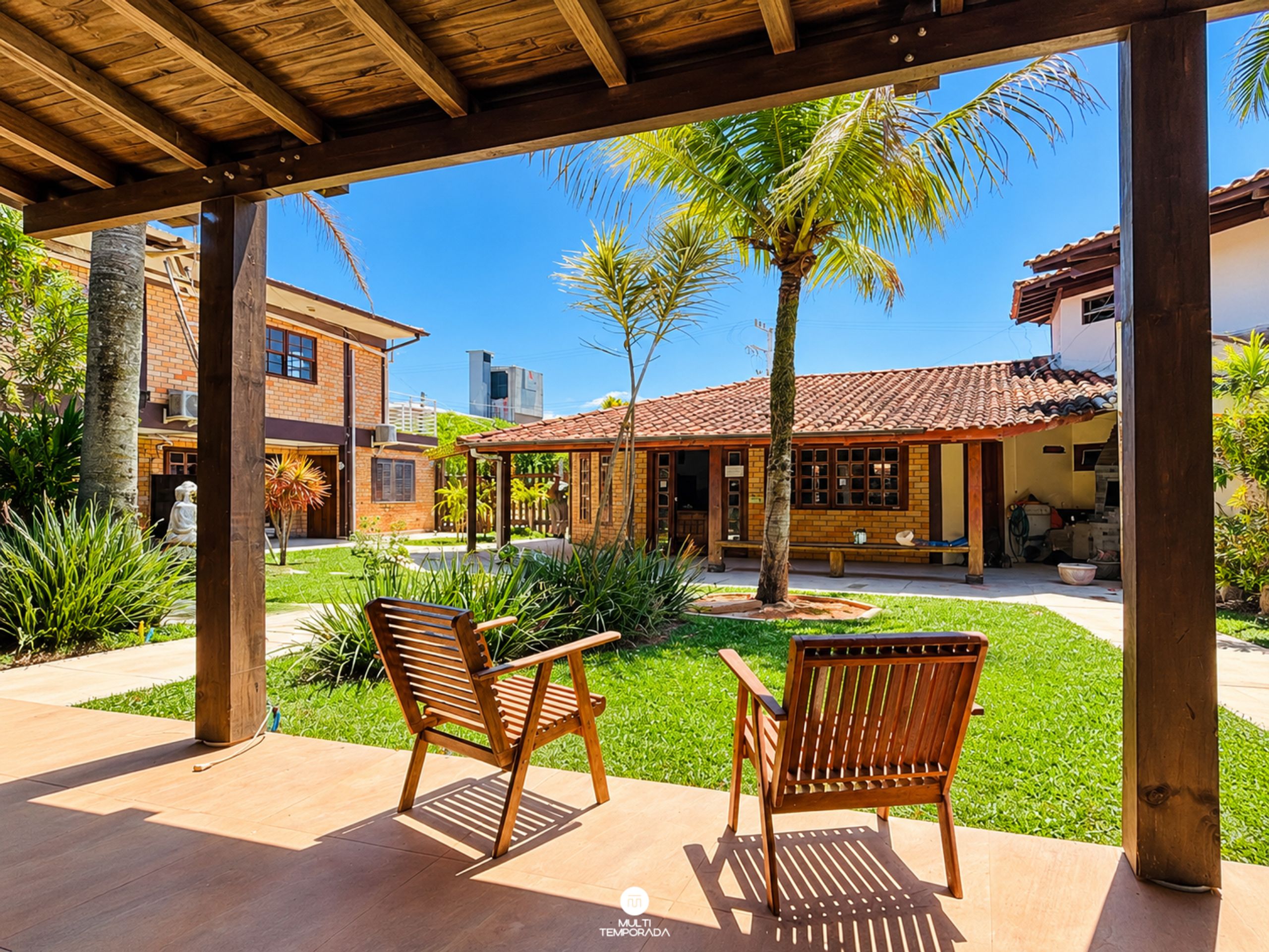 Un patio techado con dos sillas de madera, un edificio de ladrillo y una palmera al fondo.
